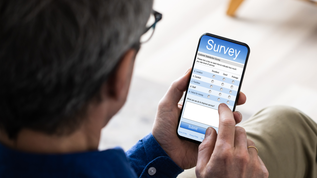 A person holding a smartphone and completing an online survey form. The screen shows multiple-choice rating options and a comment box. The person is seated indoors and tapping the phone with one hand.